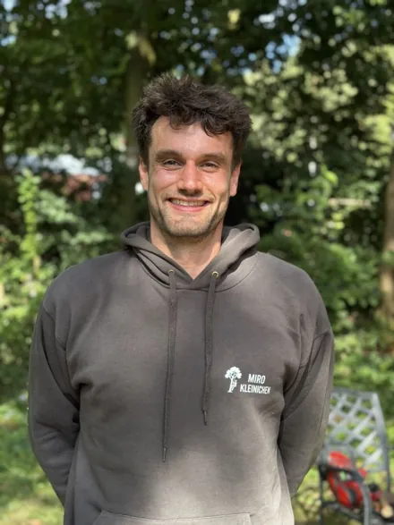 Jan Goolara Brenke im Firmenpullover vor Bäumen